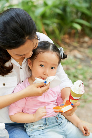 Mother using soft napkin to wipe face of little daughter after eating ice cream in park