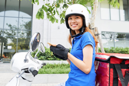 Cheerful pretty courier sitting on scooter with portable fridger behind
