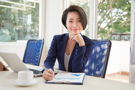 Portrait of Asian young businesswoman sitting at office desk and looking at camera while examining...