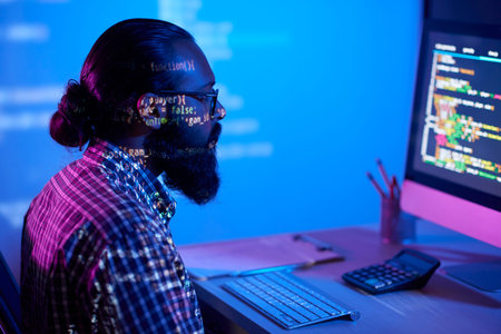 Young bearded male programmer sitting in front of computer monitor in dark, working on project in...
