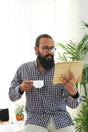 Serious bearded businessman sitting at his workplace and watching something on digital tablet while...