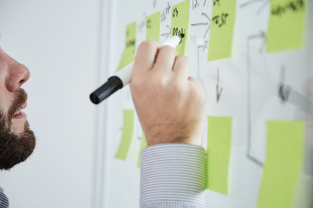 Close-up of bearded businessman writing plans with black marker on adhesive notes on the wall at...