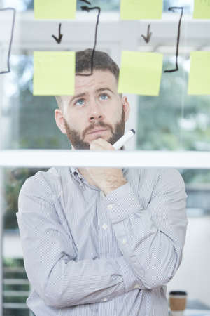 Pensive bearded businessman standing and looking at business strategy on glass wall at officeの写真素材