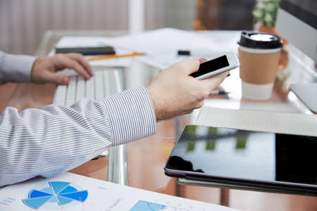 Close-up of businessman using mobile phone and communicating online while sitting at the table with tablet pc and documentsの写真素材