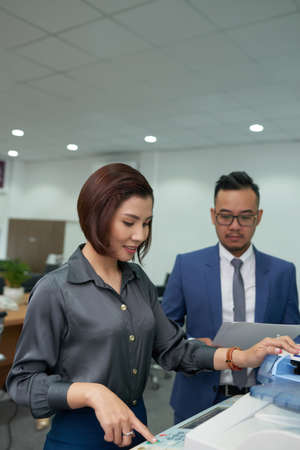 Young businesswoman making copy of document on copy machine with her colleague standing near by her and waiting for his turn at officeの写真素材