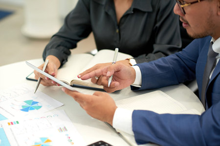 Close-up of two business partners sitting at the table pointing at digital tablet and planning new...