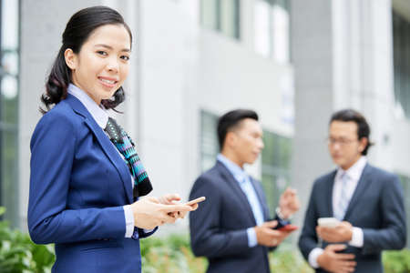 Portrait of young Asian businesswoman holding mobile phone and smiling at camera with two businessmen talking to each other in the background outdoorsの写真素材