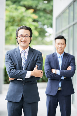 Portrait of Asian young businessman in suit and in eyeglasses standing outdoors and smiling at camera with his partner in the backgroundの写真素材