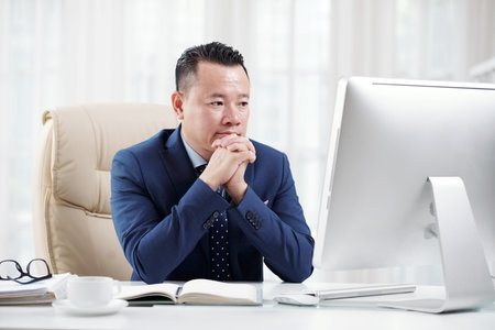 Asian mature businessman sitting at his workplace looking at computer monitor and thinking over new...
