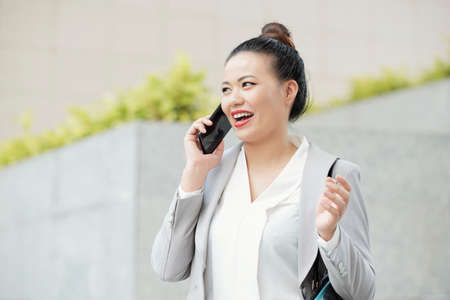 Asian young businesswoman talking on mobile phone with excitement while walking in the cityの写真素材