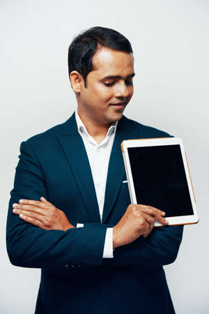 Indian businessman in suit standing with arms crossed and holding digital tablet with black empty display isolated on white backgroundの写真素材