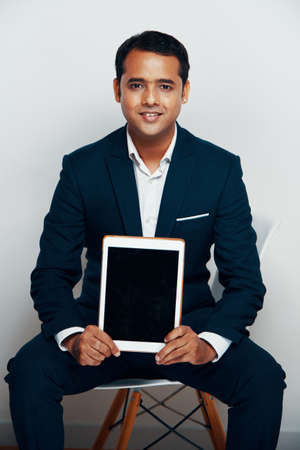 Portrait of Indian mature businessman in suit sitting on chair showing black display of digital tablet and smiling at cameraの写真素材