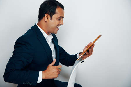 Indian mature businessman sitting on chair has online conference on digital tablet and talking to somebody with smileの写真素材