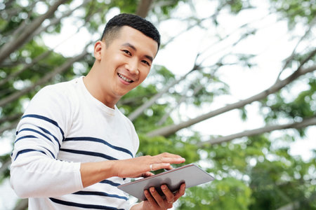 Portrait of cheerful young Vietnamese man using application on tablet computer when standing...