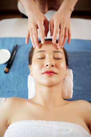 Beautician giving gentle face massage with oils to pretty young Asian patientの写真素材