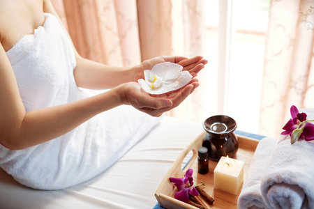Young woman sitting on bed in spa salon and holding blooming flowerの写真素材