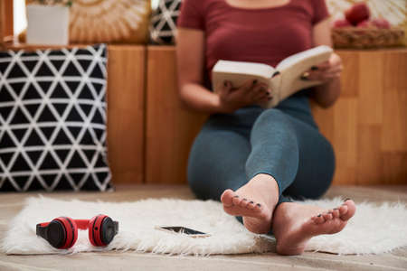 Barefoot young woman sitting of floor at home and reading good bookの写真素材