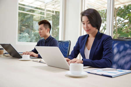 Asian business people sitting at the table and typing on laptop computers, they working online and communicating with clients during their work day at officeの写真素材