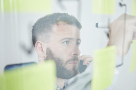 Serious young manager writing a note or reminder on the glass wall while talking on mobile phone at...