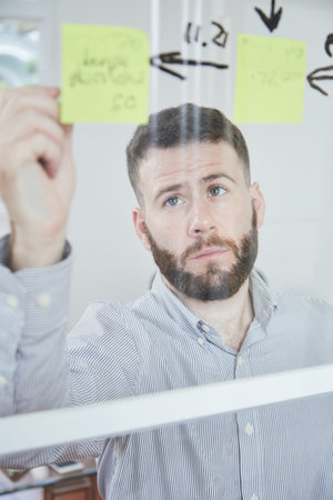 Young manager writing new ideas with marker on adhesive notes hanging on glass wall at office