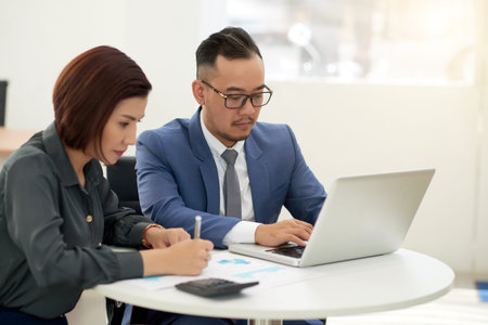 Asian young businessman in eyeglasses sitting at the table typing on laptop computer with his...