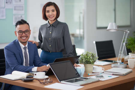 Portrait of Asian young businessman sitting at office desk typing on laptop with beautiful businesswoman standing near by him, they smiling at camera at officeの写真素材