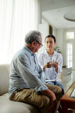 Asian young doctor sitting on sofa with senior man suggesting good pills from disease and talking about his treatment while visiting him at homeの写真素材