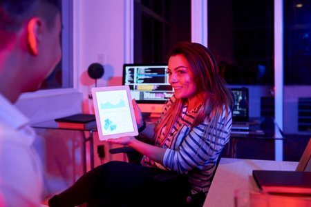 Smiling businesswoman sitting on chair holding digital tablet with financial graphs on display and explaining to her colleague in dark officeの写真素材