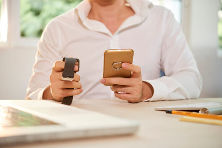Close-up of young businessman sitting at the table with wristwatch and mobile phone and checking...
