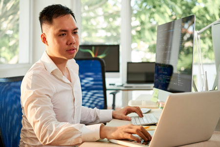 Serious Asian manager sitting at the table and configuring the software he working with computer and...