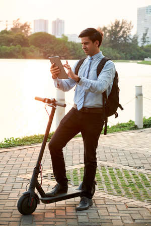 Young office worker with backpack behind his back using digital tablet and communicating online during his riding on scooter in the parkの写真素材