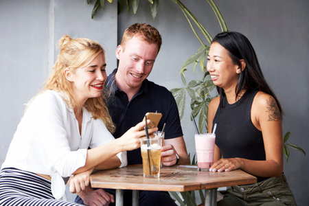 Group of friends sitting at cafe table and looking at photos in smartphone