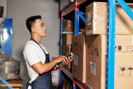 Asian young male worker standing against the shelves with cardboard boxes and checking these boxes with digital tablet in warehouseの写真素材
