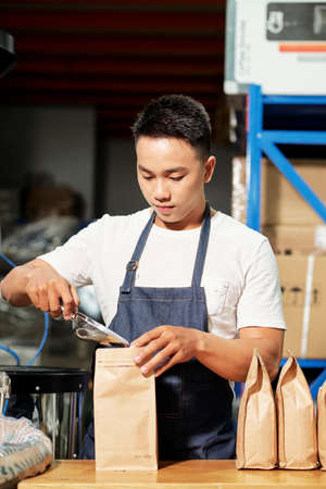 Asian man wearing apron packing freshly roasted coffee beans in craft paper bags while working in roastery houseの写真素材