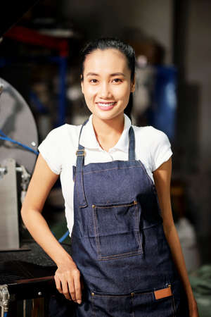 Portrait of Asian young female master standing in apron and smiling at camera while working on the factoryの写真素材