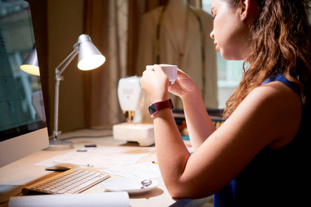 Young tired woman sitting at her workplace against the computer monitor with cup of coffee and...