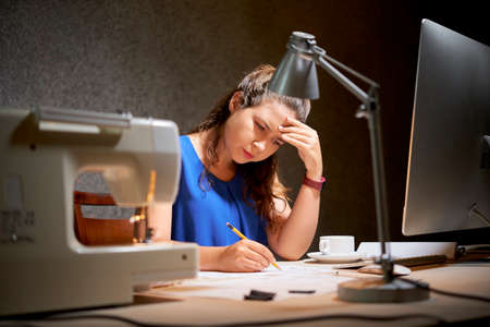 Pensive young woman thinking over new ideas and expressing it on paper at her workplace in workshopの写真素材