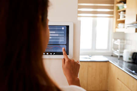Rear view of young woman making setting of her home with online smart home application which hanging on the wallの写真素材
