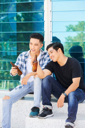 Smiling Asian friends sitting in the city discussing something and drinking beerの写真素材