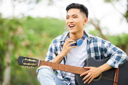 Asian smiling teenager in casual clothing and with headphones on his neck sitting with guitar in the parkの写真素材