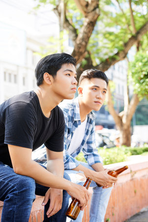 Two young Asian men sitting together outdoors with bottles of beer looking away and thinking about somethingの写真素材