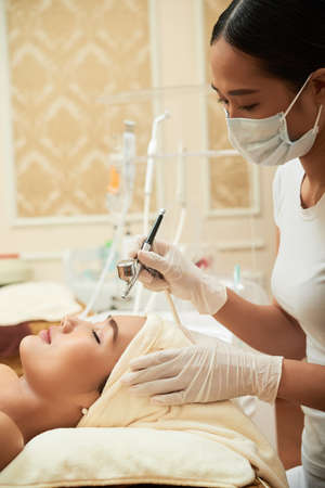 Young cosmetologist applying makeup on womans eyebrows with special equipment in cosmetological cabinetの写真素材