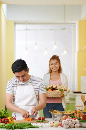 Young Asian man cutting ingredients for spaghetti dish when happy woman bringing basket of fresh...