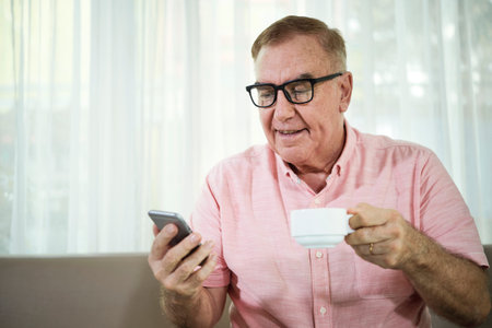 Positive senior man in glasses drinking cup of cappuccino and reading text messages in his phone