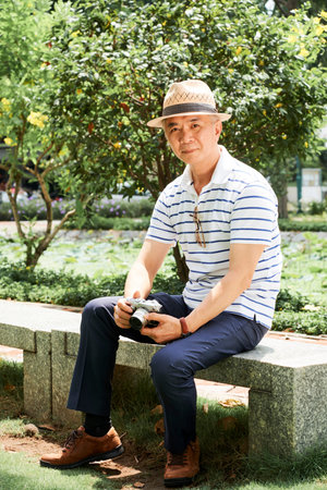 Portrait of Asian senior man in casual clothing and in hat sitting on bench with photo camera and looking at camera outdoorsの写真素材