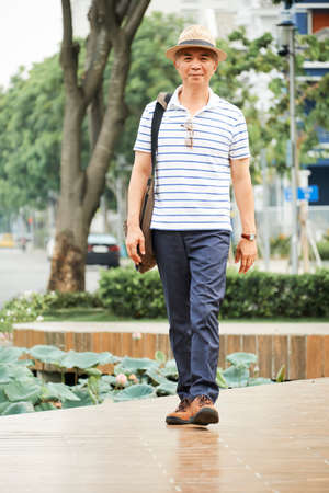 Portrait of Asian senior man in hat and in casual clothing walking in the park and looking at camera in summer dayの写真素材