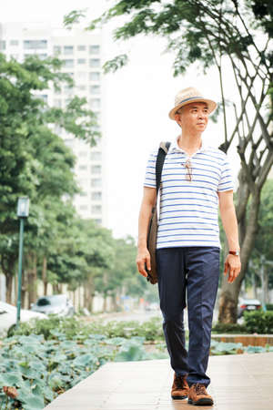 Asian senior man wearing casual clothing and hat walking in the city and enjoying the beautiful nature in summer seasonの写真素材