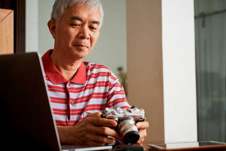 Asian senior professional sitting at the table with laptop computer and choosing the best photos from digital camera at homeの写真素材