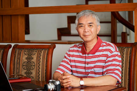 Portrait of Asian man working as a photographer he sitting at the table with laptop computer and digital camera on it and looking at cameraの写真素材
