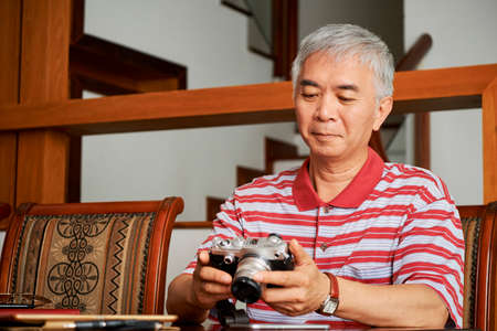 Asian mature man sitting at the table with digital camera and looking at photos after shooting dayの写真素材